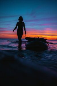 Silhouette of a woman at sunset on a beach next to a paddleboard, creating a serene and dramatic scene.